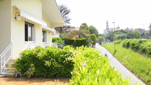 A yellow building with a covered porch, a paved walkway, and lush greenery. - Holiday Home by Lake Garda with Garden (Lazise)