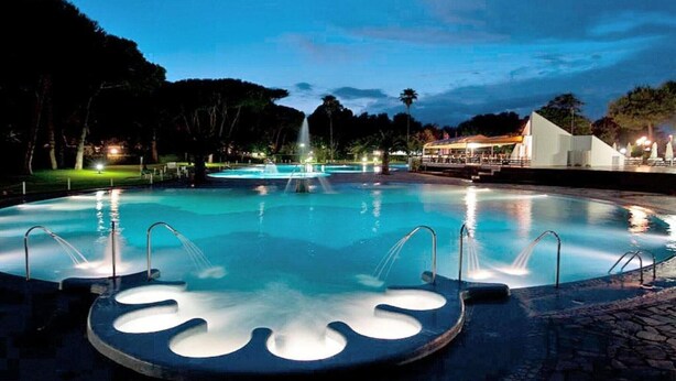 A nighttime swimming pool with water jets, surrounded by trees and a building with a triangular roof. - Chalet in Baia Domizia with Private Beach (Baia Domizia (CE))