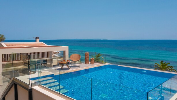 Una piscina infinita moderna con agua azul cristalina, con vistas al mar turquesa y un edificio blanco con tejado de tejas rojas.