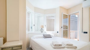 A bedroom with a bed, a nightstand, a lamp, a window with shutters, and a view of the outside.