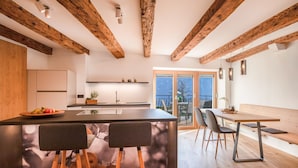 Une cuisine moderne avec des poutres en bois, un îlot central doté de tabourets, un coin repas avec un banc et une vue dégagée sur l’extérieur grâce à une porte vitrée.
