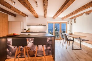 A modern kitchen with wooden beams, a central island with stools, a dining area with a bench, and a view through a glass door.