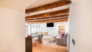 Une cuisine moderne avec des poutres en bois, un coin repas avec des tabourets de bar, un téléviseur à écran plat et un canapé gris dans un espace de vie lumineux.