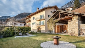 Une maison à deux étages avec une façade en pierre, une terrasse en bois et un foyer circulaire dans le jardin.