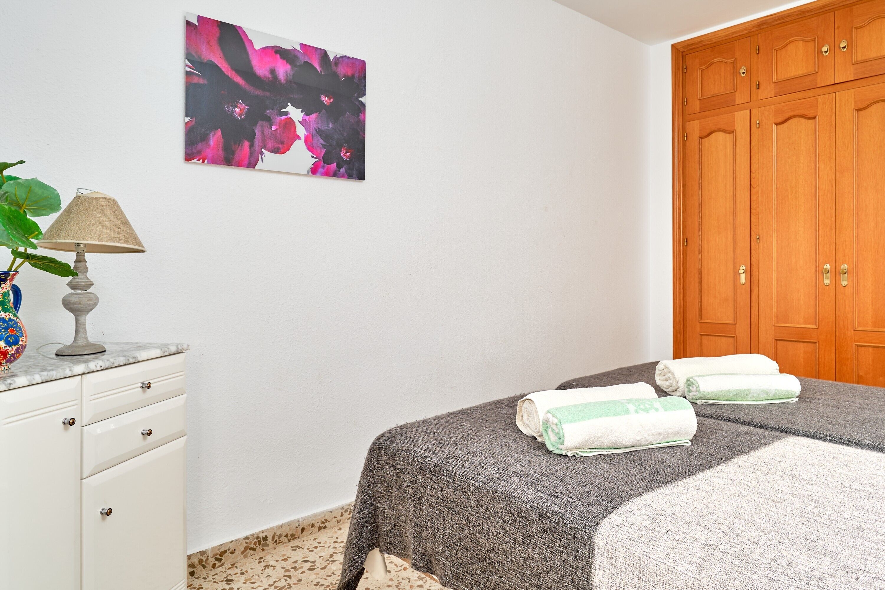 Un dormitorio con cama, armario de madera, escritorio con lámpara y un cuadro en la pared.