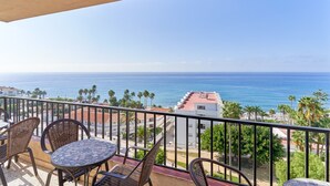 Un balcon agrémenté de chaises et d’une table en osier, offrant une vue sur la côte, avec des bâtiments et des palmiers.