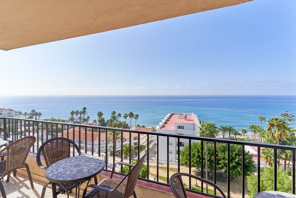 A balcony with wicker chairs and a table, overlooking a coastal view with buildings and palm trees.