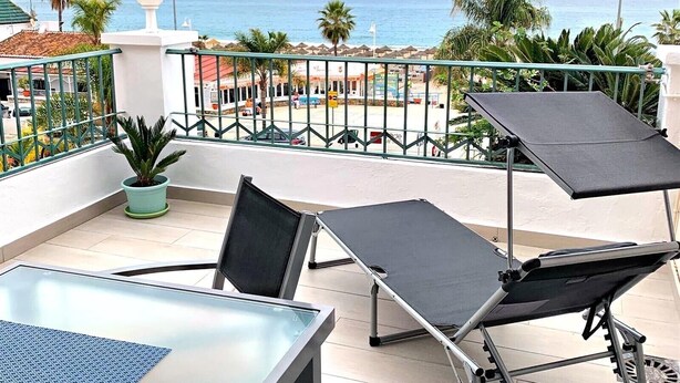 Ein Balkon mit Tisch und Stühlen, einem Blumentopf und Blick auf Strand und Meer.