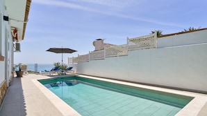 Una piscina rectangular amb aigua cristal·lina, envoltada per una paret blanca amb una tanca decorativa, i vista al mar.