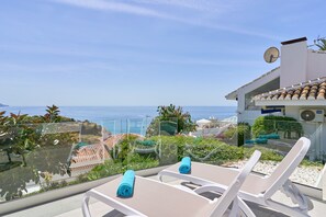 A balcony with two white lounge chairs, a glass barrier, and a view of the sea and buildings.