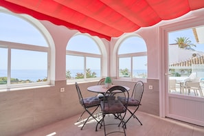 Eine sonnendurchflutete Terrasse mit Tisch und Stühlen, großen Bogenfenstern und Meerblick.