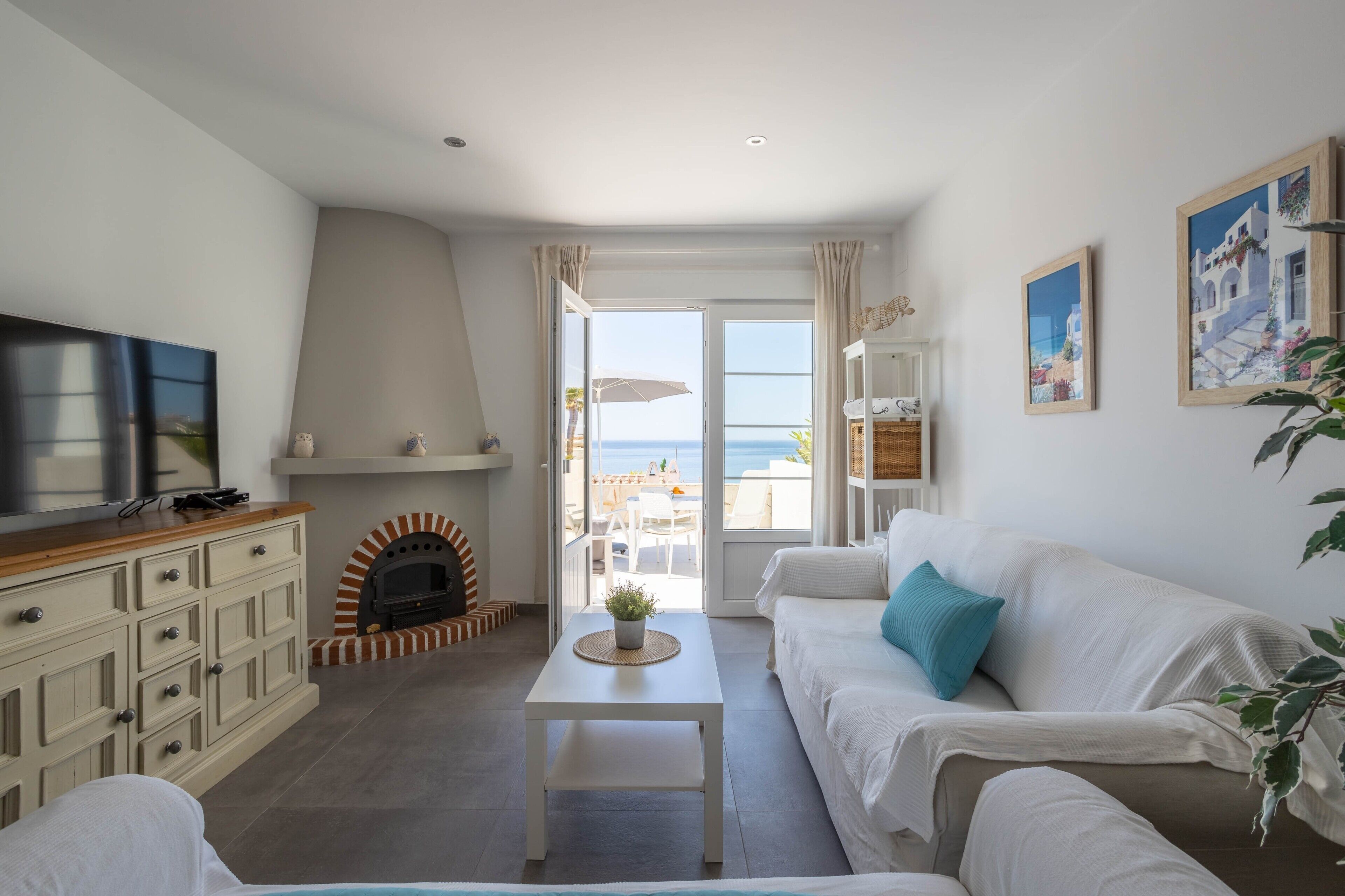 Een woonkamer met een open haard, een televisie, een bank en uitzicht op de zee door grote ramen.