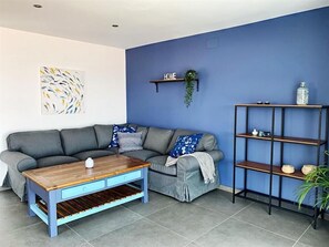 A living room with a grey sectional sofa, a wooden coffee table with drawers, a blue wall, a 'HOME' sign, a plant, and a black metal shelving unit.