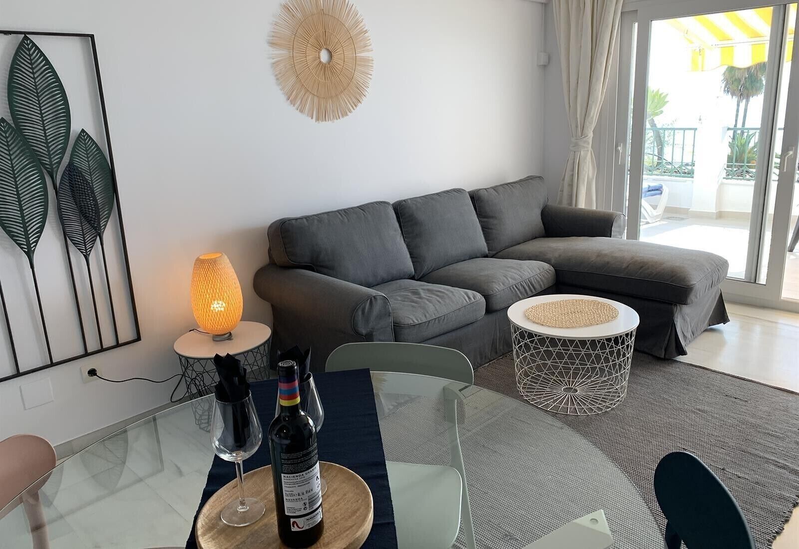 A modern living room with a grey sofa, a glass dining table with a bottle of wine, and a decorative wall piece.