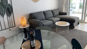 Een moderne woonkamer met een grijze bank, een glazen eettafel met een fles wijn, en een decoratief wandobject.