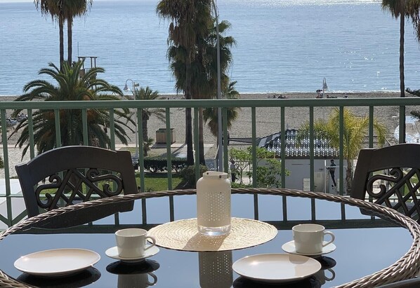 Une table ronde entourée de chaises, une carafe et des verres, le tout sur un balcon donnant sur la mer.
