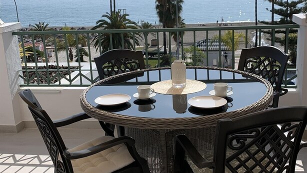 Een ronde tafel met stoelen, een glazen karaf en witte bekers op een balkon met uitzicht op zee.