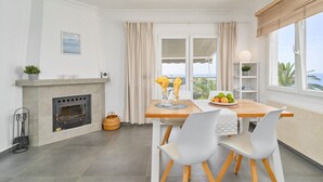 Une cuisine moderne avec une cheminée, une table à manger avec des chaises et une vue sur la mer grâce à de grandes fenêtres.
