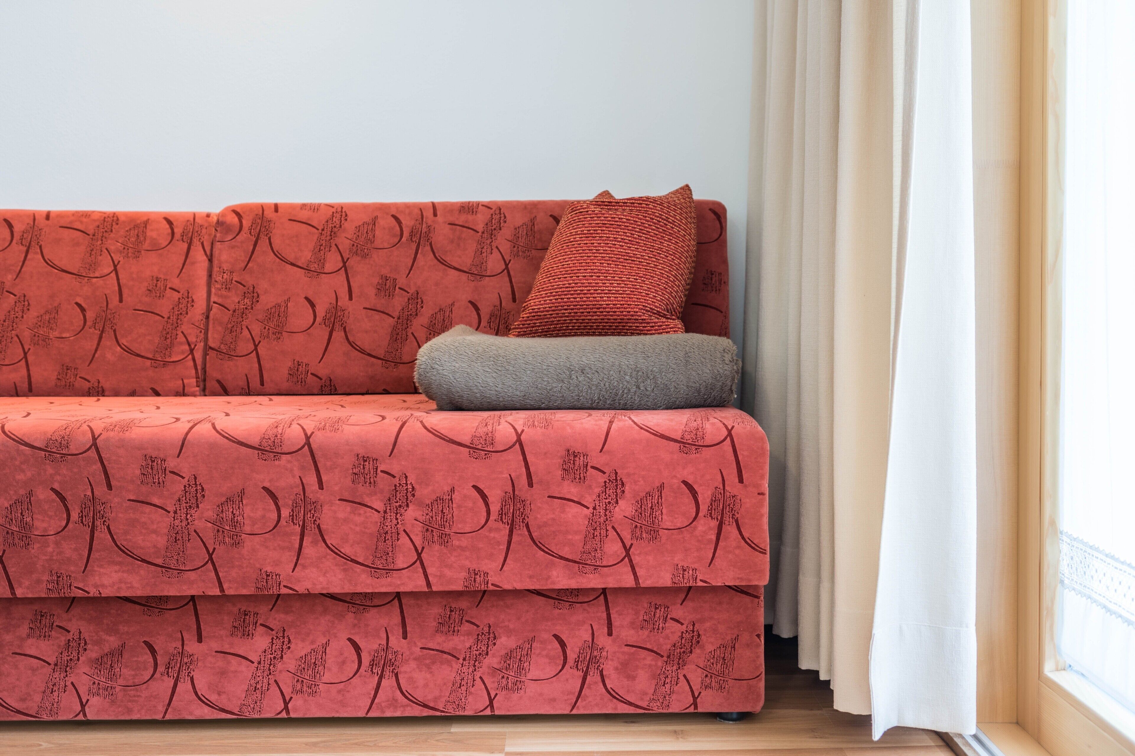 A red sofa with a patterned cover, a grey cushion, and a white curtain partially drawn.