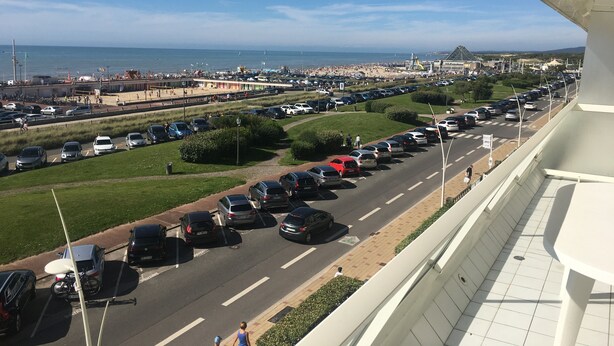 Une vue d’une route côtière bordée de voitures garées, d’une plage et d’un stade situé au loin.