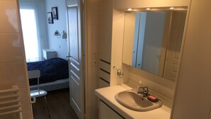 Une salle de bain moderne avec un lavabo blanc, un miroir et un meuble. Dans la pièce adjacente, on trouve un lit recouvert d’une couverture bleue.