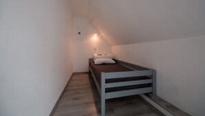 Une petite pièce étroite, avec un lit simple, un luminaire fixé au mur et une porte dotée d’une fenêtre ronde.