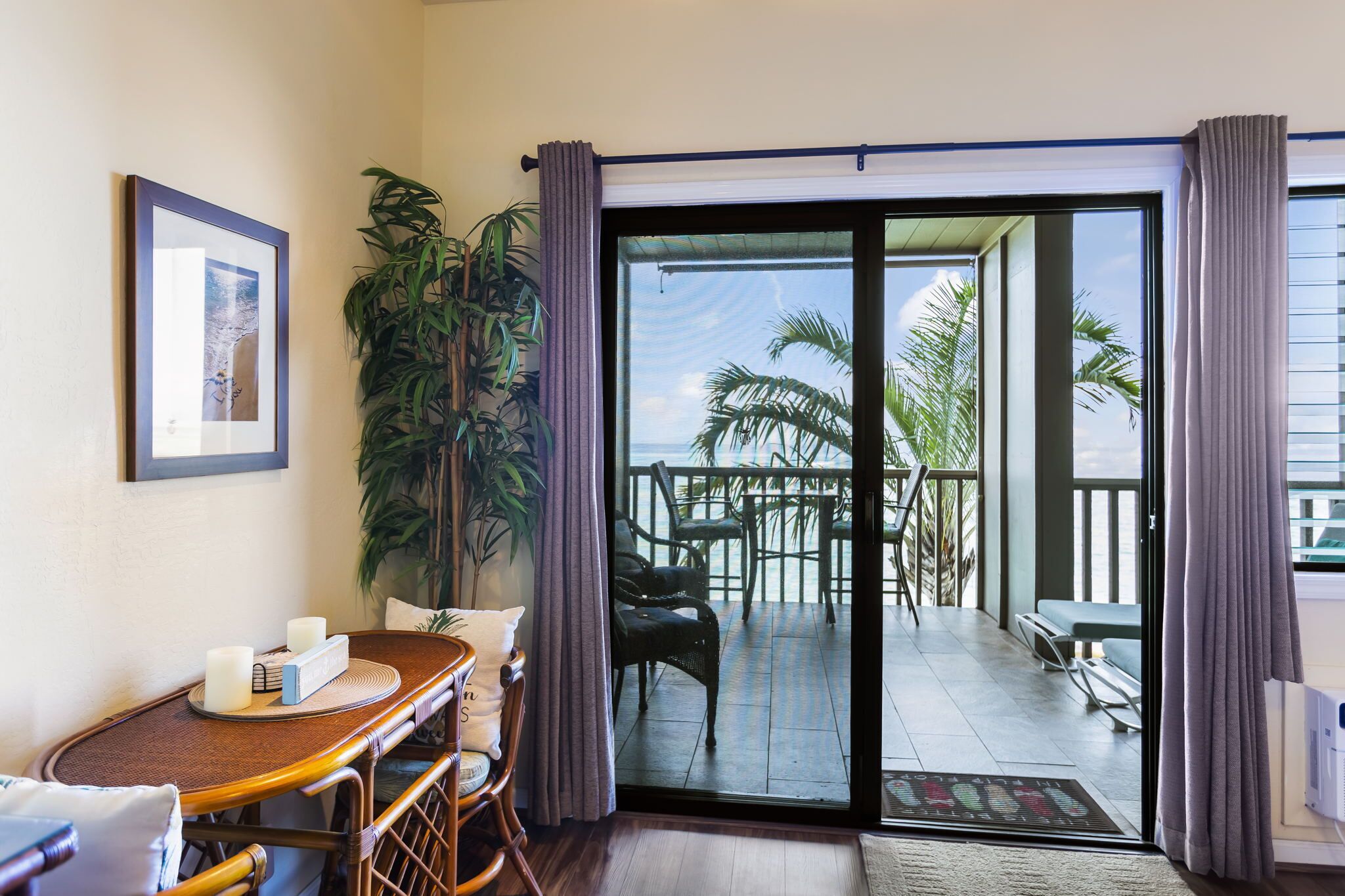A room with a wooden table, chairs, a potted plant, and a sliding glass door leading to an outdoor deck with chairs and a view of palm trees.