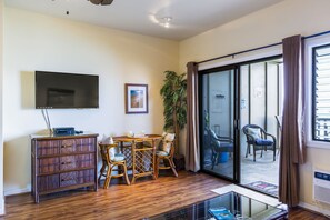 A living room with a flat-screen TV, a wooden cabinet, a dining table with chairs, a potted plant, and a sliding glass door leading to an outdoor area.