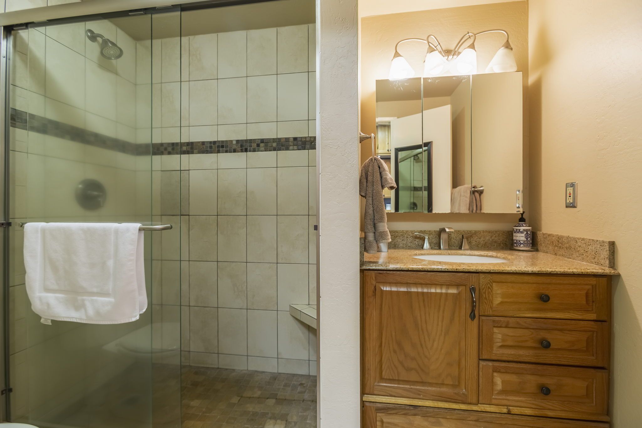 A bathroom with a glass shower enclosure, a white towel hanging on a rack, a wooden vanity with a sink, and a mirror with lights above it.