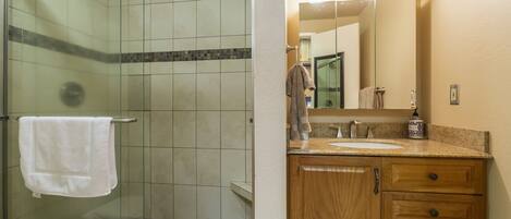 A bathroom with a glass shower enclosure, a white towel hanging on a rack, a wooden vanity with a sink, and a mirror with lights above it.