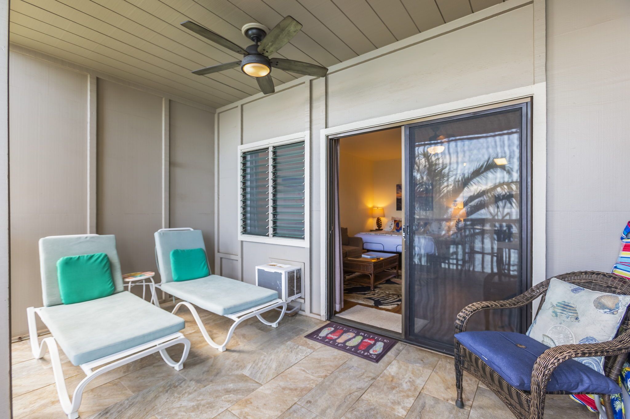 A covered patio with two lounge chairs, a wicker chair, and a sliding glass door leading to an interior space with a table and chairs.