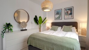 A modern bedroom with a large bed, a round mirror, two wall-mounted pictures, and a bedside table with a lamp.