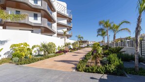 A modern multi-story building with balconies, surrounded by a landscaped garden with various plants and palm trees.