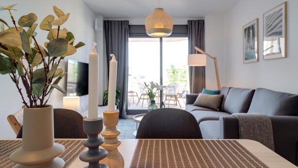 A modern living room with a sofa, a dining table with placemats, a potted plant, and a tall candle.
