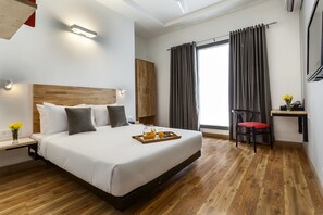 Une chambre d’hôtel avec un grand lit, une tête de lit en bois, des tables de chevet avec des lampes, un bureau avec une chaise et une fenêtre avec des rideaux.