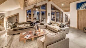 Ein geräumiges Wohnzimmer mit hohen Decken und Holzbalken, ausgestattet mit einer großen Couchgarnitur, einem Couchtisch mit Büchern und dekorativen Gegenständen, und Blick ins Innere durch große Glasüren.