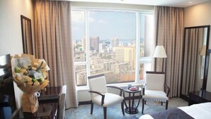 Quarto de hotel com vista para a cidade, uma cama, duas cadeiras, uma mesa pequena, um abajur e um arranjo de flores.