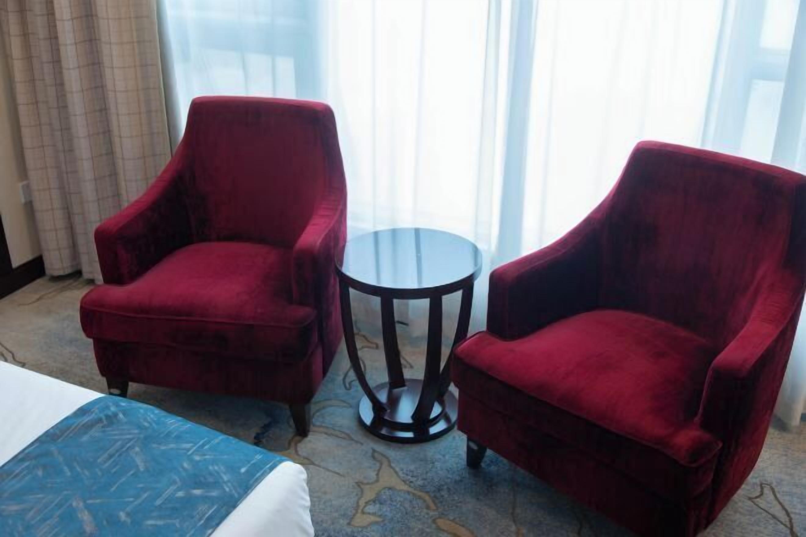 A hotel room with two red armchairs, a small round table, and a bed with a blue and white patterned bedspread.