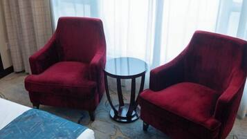 A hotel room with two red armchairs, a small round table, and a bed with a blue and white patterned bedspread.