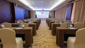 Un salón de conferencias con filas de mesas y sillas, una pantalla de proyector al frente y piso alfombrado.