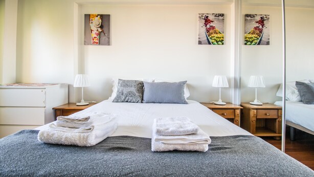 Una cama bien hecha con dos toallas dobladas, un edredón gris y dos mesitas de noche con lámparas. Se observan dos cuadros colgados en la pared y un aparador blanco.