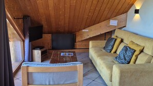 Une pièce chaleureuse avec un plafond en bois, un canapé agrémenté de coussins à motifs, une table en bois sur laquelle repose un magazine et un téléviseur posé sur un meuble.