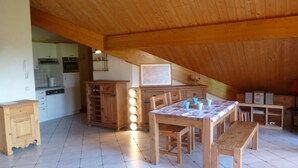 Une pièce mansardée spacieuse avec un plafond en bois, une table à manger avec des chaises, un coin cuisine avec des placards et un lave-vaisselle, et une bibliothèque.