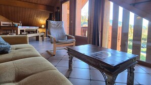 Une pièce chaleureuse avec un plafond en bois, un canapé confortable, un fauteuil à bascule, une table en bois et de grandes fenêtres donnant sur l’extérieur.