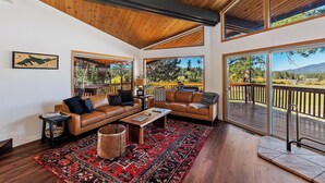 A spacious living room with a wooden ceiling, large windows, a leather sofa, a coffee table, and a red patterned rug.