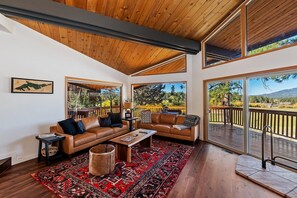 A spacious living room with a high ceiling, large windows, and a view of a deck and trees.