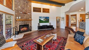 A spacious living room with a stone fireplace, a large flat-screen TV mounted on the wall, and a cozy seating area with a red patterned rug.