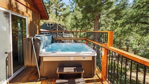 Un bain à remous sur une terrasse en bois, avec une porte vitrée donnant sur une pièce, le tout entouré d’arbres.