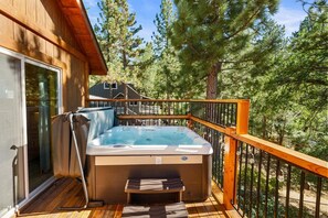Un bain à remous sur une terrasse en bois, avec une porte vitrée donnant sur une pièce, le tout entouré d’arbres.