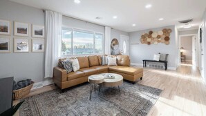 A modern living room with a brown leather sectional sofa, a round coffee table, and a patterned rug. There are framed pictures on the wall, a large window with curtains, and a wall-mounted basket. - Denver Retreat • Hot tub+Firepit+Games • Fast Wifi (DENVER)
